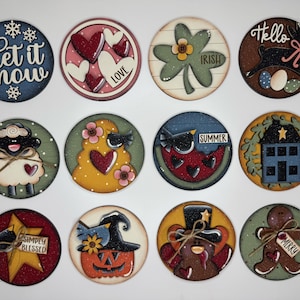May include: A collection of twelve round wooden ornaments with various designs. The ornaments feature seasonal themes, including a snowflake, hearts, a shamrock, a bunny, a sheep, a bird, a watermelon, a house, a star, a pumpkin, a bear, and a gingerbread man. Each ornament has a unique color palette and text.