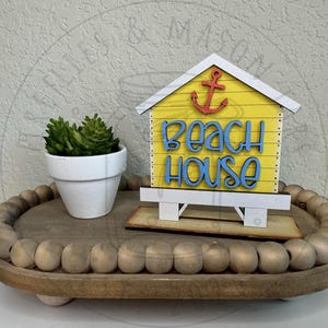 以下が含まれることがあります： 赤い錨と青い「beach house」の文字が描かれた黄色い木製のビーチハウス型サインが、ビーズの縁取りがある木製トレイの上に置かれています。隣には緑色の多肉植物が入った小さな白い鉢があります。サインには白い屋根と土台が付いています。