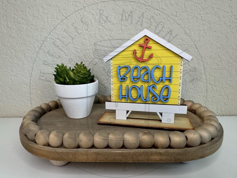 以下が含まれることがあります： 赤い錨と青い「beach house」の文字が描かれた黄色い木製のビーチハウス型サインが、ビーズの縁取りがある木製トレイの上に置かれています。隣には緑色の多肉植物が入った小さな白い鉢があります。サインには白い屋根と土台が付いています。