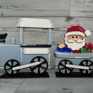 May include: Wooden Christmas train set with a light blue engine and a car carrying Santa Claus. The engine has black wheels and a white roof. Santa is wearing a red hat and holding gifts. The set is on a wooden base.