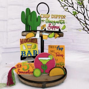 May include: A two-tiered wooden serving tray with a black metal stand. The tray is decorated with various wooden signs and cutouts with a summery, margarita-themed design. The signs include phrases like "Chip Dippin' + Margarita Sippin'," "Margarita Bar," and "It's 5 O'Clock Somewhere." There are also cactus cutouts, margarita glass cutouts, and taco cutouts.