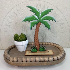 以下が含まれることがあります： 緑色の葉と茶色の質感のある幹を持つ木製のヤシの木の切り抜きが、長方形の台座の上に立っています。その隣には、緑色の多肉植物が入った小さな白い陶器の鉢があり、すべてビーズの縁取りが付いた楕円形の木製トレイの上に配置されています。