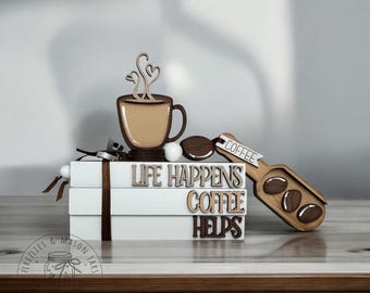 May include: A stack of three white books with the words "Life Happens Coffee Helps" on them. A brown coffee cup with steam coming out of it is on top of the books. A brown wooden coffee scoop with coffee beans on it is next to the books.