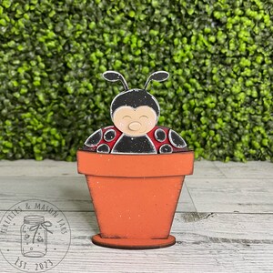 Puede incluir: Una figurita de mariquita de madera pintada de negro y rojo, sentada dentro de una peque&ntilde;a maceta de flores naranja. La mariquita tiene una cara blanca con ojos negros y una peque&ntilde;a nariz rosa.