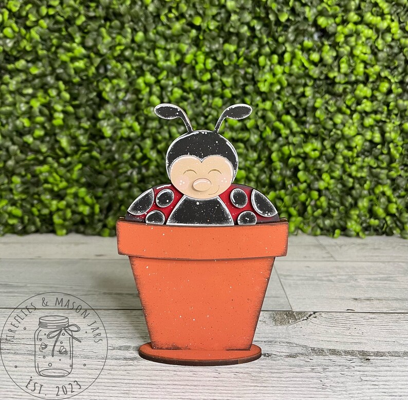 Puede incluir: Una figurita de mariquita de madera pintada de negro y rojo, sentada dentro de una peque&ntilde;a maceta de flores naranja. La mariquita tiene una cara blanca con ojos negros y una peque&ntilde;a nariz rosa.
