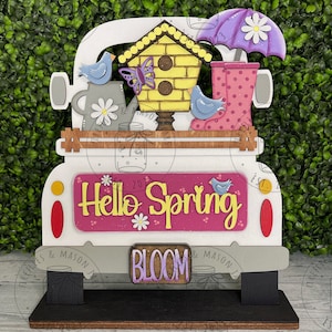 May include: A white wooden truck with a pink and yellow "Hello Spring" sign on the back. The truck is carrying a birdhouse, a butterfly, two blue birds, a watering can, a purple umbrella, and pink rain boots. The truck is on a black stand with the word "BLOOM" on the front.