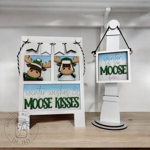 May include: White wooden easel with a winter theme. The easel has two moose illustrations with the text "winter wishes & moose kisses". A separate white wooden sign with a black string hangs from the top. The sign has the text "winter is so moose fun".