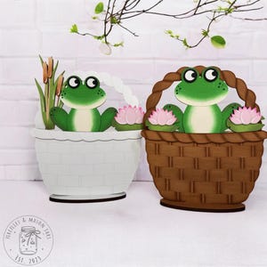May include: Two wooden frog figurines sitting in baskets. One basket is white and the other is brown. Both frogs are green with black eyes and are holding pink lily pads.