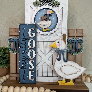 May include: Wooden farmhouse-style decorative piece featuring a white barn door with a goose illustration, a sign that reads "hey silly goose", and a wooden goose. The piece is adorned with blue bows and greenery, and a string of wooden beads.
