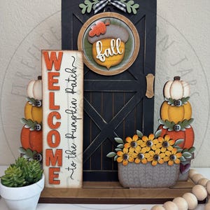 May include: A decorative wooden display featuring a black barn door with a fall acorn sign, a vertical sign reading WELCOME to the Pumpkin Patch, a stack of three pumpkins, a basket of yellow flowers, and a small green succulent in a white pot.