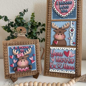 May include: Two wooden decorative signs with moose and heart designs. One sign is square and the other is rectangular. The signs have the phrases "It Moose Be" and "I Moose Love You" and "You Moose Be Mine". The moose have pink bows and hearts.