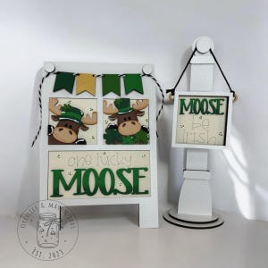 May include: A white wooden sign with a green shamrock and moose design. The sign reads "one lucky moose" and has a banner with green and gold flags. A smaller white sign with a green shamrock and moose design reads "Moose be Irish".