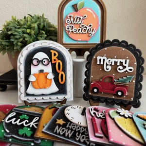 May include: A collection of decorative wooden signs with various designs. One sign says "Just Peachy" with a peach illustration. Another features a ghost holding a pumpkin with "BOO". Other signs say "merry", "luck", and "Happy New Year".