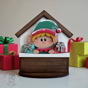 May include: Wooden Christmas elf decoration in a house-shaped design. The elf wears a green hat with white speckles and a red trim. The elf is holding two wrapped gifts. The house has a brown roof and a white facade. The base of the house is brown.