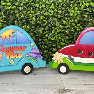 May include: Two wooden car shaped ornaments, one painted blue with the words "Summer Vibes" and the other painted red and green to resemble a watermelon.