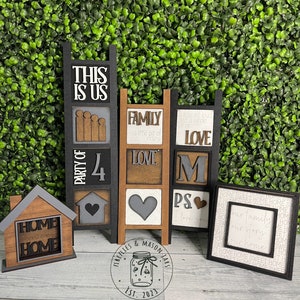 May include: A set of five wooden signs with black frames and white lettering. The signs are arranged in a vertical stack and feature the phrases "This is Us", "Party of 4", "Family", "Love", and "PS I love you". The signs are decorated with gray and brown hearts. The signs are sitting on a wooden surface with a green background.