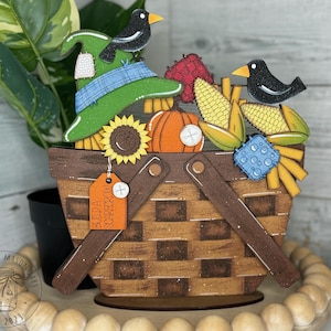 May include: A wooden craft project depicting a fall harvest scene. A brown wicker basket is filled with corn, pumpkins, and a scarecrow hat. Two black birds are perched on the basket. A tag on the basket reads "Build a Scarecrow".