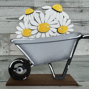 May include: A gray wooden wheelbarrow filled with white and yellow daisies. The wheelbarrow is on a brown wooden base.