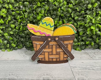 May include: A wooden cutout of a brown picnic basket with a yellow sombrero and a yellow taco inside. The basket has a brown handle and a white and brown woven pattern.