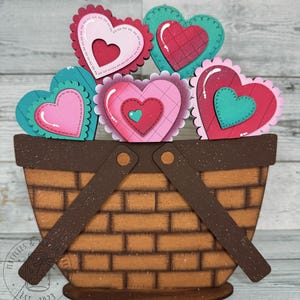 May include: A wooden basket filled with five colorful hearts. The hearts are decorated with different patterns and textures. The basket is brown with a brown handle. The basket is sitting on a wooden surface.