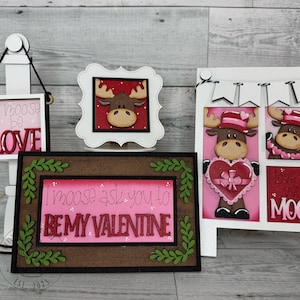 May include: Three wooden signs with a Valentine's Day moose theme. The first sign says "I moose be love". The second sign says "I moose ask you to be my Valentine". The third sign has two moose, one holding a heart and the other saying "I love you very moose".