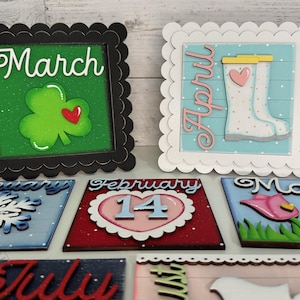 May include: Twelve wooden signs with scalloped edges, each representing a month of the year. The signs feature various illustrations and text, including a shamrock for March, rain boots for April, a snowflake for January, a heart with the number 14 for February, flowers for May, and the word "July" for July.