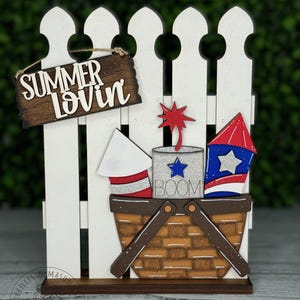May include: A white picket fence with a wooden sign that says "Summer Lovin'" hanging from it. There is a red, white, and blue firework in a brown wicker basket with the word "BOOM" written on it.