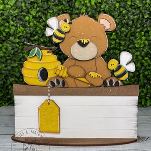 May include: A wooden sign with a brown bear, a beehive, and two bees. The sign has a white background with a brown border and a brown base. The sign has a yellow tag that says "Sweet Treats".