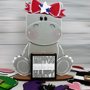 May include: A gray hippo-shaped decoration with a red, white, and blue bow. The hippo holds a sign that reads "Hippos make me HAPPY." Additional decorative cutouts are scattered around the base, including a witch hat and hearts.