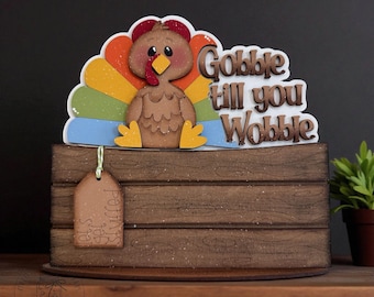 以下が含まれることがあります： カラフルな尾羽と「Gobble till you Wobble」のフレーズが特徴の、木製の感謝祭の装飾。 ターキーは、「Let's get stuffed」と書かれた小さなタグが付いた木製の箱の上に座っています。