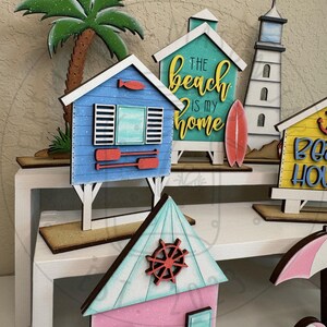 以下が含まれることがあります： ヤシの木、赤いオールが付いた青いビーチハウス、THE beach IS MY homeという文字が入ったターコイズブルーの家、灯台、黄色いビーチハウス、舵輪が付いたピンクの家など、木製のビーチをテーマにした装飾品のコレクション。