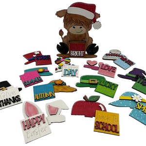 May include: A set of 18 wooden holiday and seasonal signs featuring various themes, including "Give Thanks", "Happy Easter", "Hello Summer", "Hello Autumn", "Winter Wishes", "Spring Flowers", "Back to School", "Trick or Treat", "Lucky Wishes", "Love You More", and "Day". The signs are decorated with glitter, paint, and various embellishments. A wooden Highland cow figurine wearing a Santa hat sits on top of a sign that reads "BRIGHT".