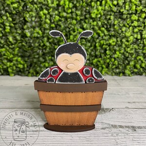 Puede incluir: Una figurita de mariquita de madera pintada de negro y rojo, sentada dentro de un barril de madera marr&oacute;n. El barril est&aacute; sobre una base de madera.