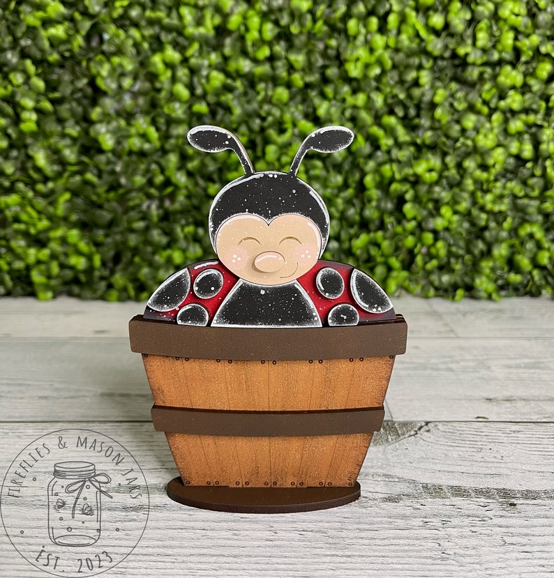 Puede incluir: Una figurita de mariquita de madera pintada de negro y rojo, sentada dentro de un barril de madera marr&oacute;n. El barril est&aacute; sobre una base de madera.