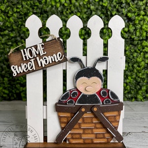 Puede incluir: Un letrero de madera con la frase "Home sweet home" con una mariquita en una cesta, colgado de una valla blanca de madera.
