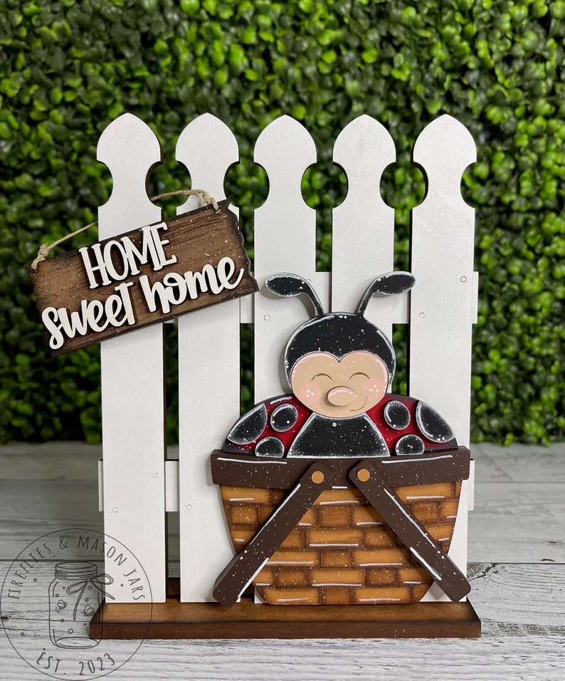 Puede incluir: Un letrero de madera con la frase "Home sweet home" con una mariquita en una cesta, colgado de una valla blanca de madera.