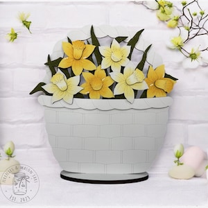 May include: A white wooden basket filled with yellow daffodils. The basket has a woven texture and sits on a wooden base. The daffodils are arranged in a bouquet and have green leaves.