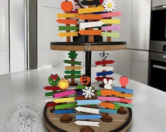 May include: A two-tiered wooden tiered tray with a black metal stand. The top tier has a variety of colourful wooden signs with autumn and winter themes. The signs have text such as "Gobble Drive", "Pumpkin Pie St", "Turkey Trail", "Firecracker Ct", "Snowflake Lane", and "Frosty Freeway Winter Way".