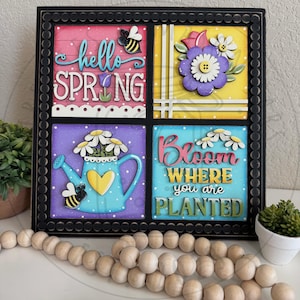 May include: A square, framed wall art piece with a black border. The art is divided into four colorful squares, each featuring spring-themed designs, including flowers, bees, and the phrases "hello SPRING" and "Bloom where you are planted."