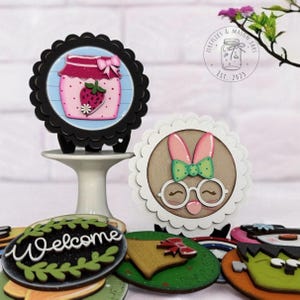 May include: A set of five wooden round signs with various designs. The signs feature a jar of strawberry jam, a bunny with glasses, a welcome sign with greenery, a bell, and a snowman. The signs are all painted in bright colors and have a whimsical feel.