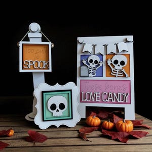 May include: Halloween-themed wooden decorations featuring a skull, skeletons, and pumpkins. One sign reads "Too cute to spook," and another says "These bones love candy." The decorations are white, with orange, blue, and pink accents, and are set on a wooden surface.
