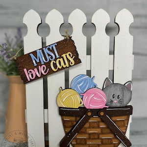 May include: A wooden cat-themed decorative piece featuring a white picket fence, a basket with yarn balls, and a gray cat head. The sign reads "MUST love cats" in colorful letters. The basket holds yellow, blue, and pink yarn.
