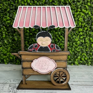 Puede incluir: Un puesto de mercado de madera con forma de mariquita, con un toldo rosa y un letrero que dice "Market stand".