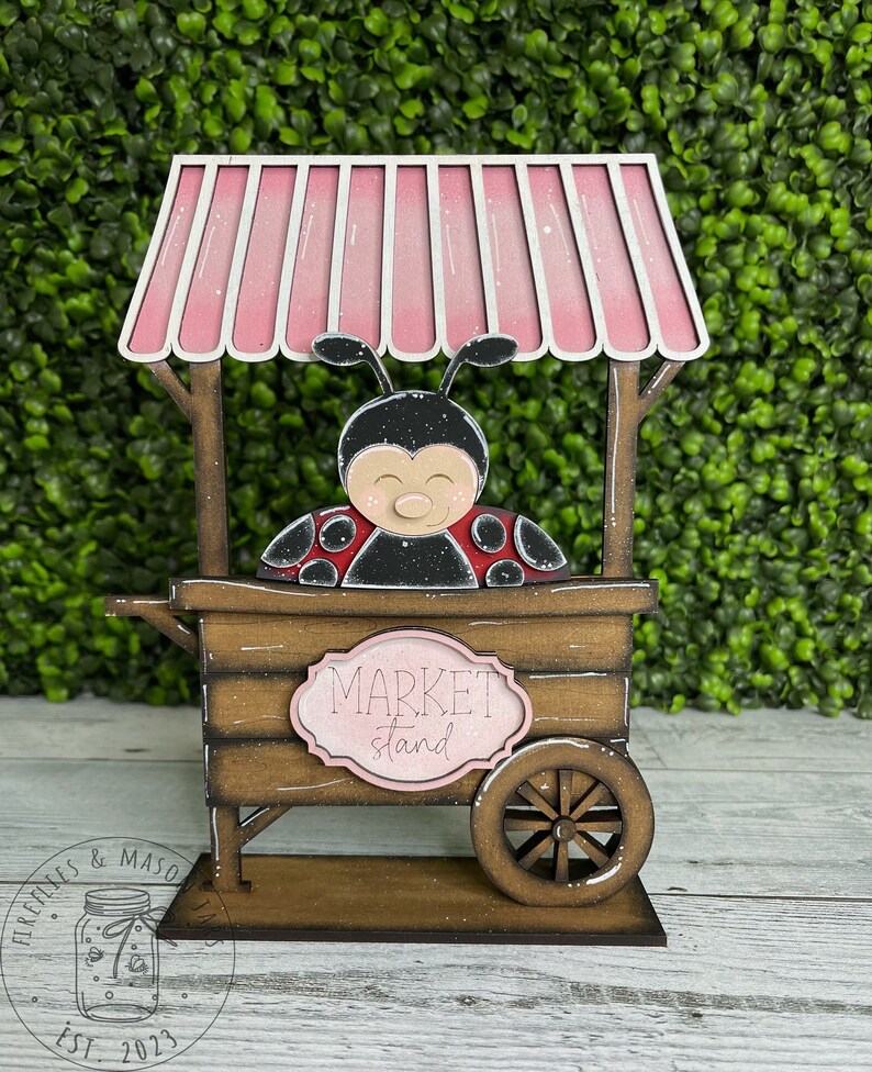 Puede incluir: Un puesto de mercado de madera con forma de mariquita, con un toldo rosa y un letrero que dice "Market stand".