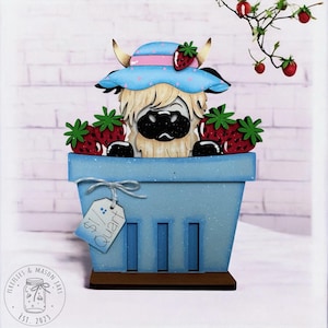 May include: A wooden decorative piece featuring a Highland cow wearing a blue hat with a pink band and strawberry accents, peeking out of a blue basket filled with strawberries. The basket has a price tag that reads "$1/ Quart".