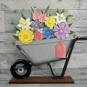 May include: A wooden wheelbarrow filled with a colorful bouquet of flowers. The flowers are painted in shades of pink, yellow, purple, and white. The wheelbarrow is painted gray and black and has a red tag that says "Fresh Cut Flowers".
