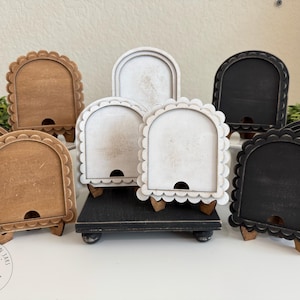 May include: A collection of wooden arched display stands in various colors: brown, white, and black. Each stand has a scalloped edge and a small hole at the bottom. Some stands are on a black platform. The stands are for displaying items.