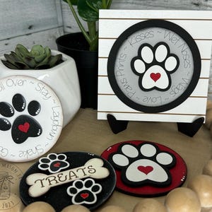 May include: Four wooden signs with paw prints and the text "Dog ~ Sit ~ Good ~ Shake ~ Laydown ~ Roll Over ~ Stay", "Love ~ Paws ~ Prints ~ Kisses ~ Wet Noses ~ Wags", "TREATS", and a paw print with a heart. The signs are on a wooden surface with a white background.