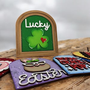 May include: Four wooden signs with different themes. The first sign is green with a shamrock and the word "Lucky". The second sign is purple with a bunny wearing glasses and the word "Easter". The third sign is blue with stars and the words "Boom Chicka Pop". The fourth sign is red, white, and blue with a cowboy hat and the words "Happy 4th".