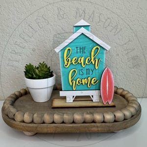 以下が含まれることがあります： ターコイズブルーのファサードに黄色の文字で「THE beach IS MY home」と書かれた木製のビーチハウス型看板と、小さなピンクのサーフボードの切り抜き。白い鉢植えの多肉植物と木製ビーズが飾られた木製トレイの上に置かれています。